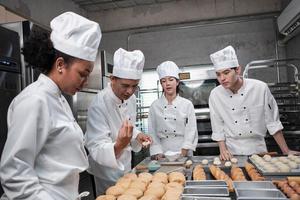 equipo gourmet profesional multirracial, cuatro chefs con uniformes de cocina blancos y delantales amasan masa de pastelería y harina, preparan pan y comida de panadería, hornean en el horno en la cocina del restaurante de acero inoxidable. foto