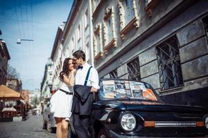 pareja joven, mujer y hombre guapo posando cerca de un auto retro negro foto