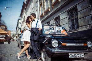 pareja joven, mujer y hombre guapo posando cerca de un auto retro negro foto