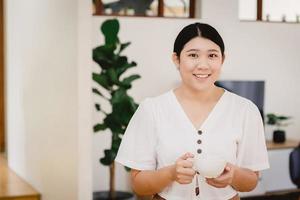 una linda adolescente asiática maneja una taza y sonríe para hacer dieta bebiendo té de hierbas o una bebida saludable en casa.