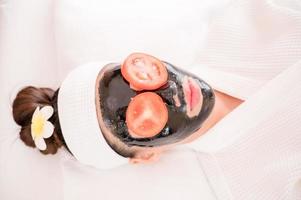 una hermosa mujer asiática usa lodo de spa y rodaja de tomate para el tratamiento facial foto