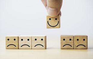 Hand holding Smile face print screen on wooden cube block among sadness face for customer service evaluation and emotion mindset concept. photo