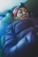 una mujer con un faro yace en un saco de dormir en una carpa y sostiene un teléfono inteligente foto
