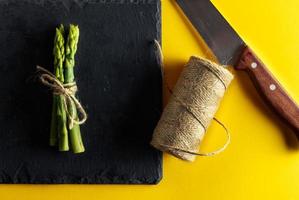 Asparagus. Bunch of fresh asparagus tied on a plate of slate with a knife and a roll of string on yellow background. Healthy vegetarian food. Horizontal image. photo