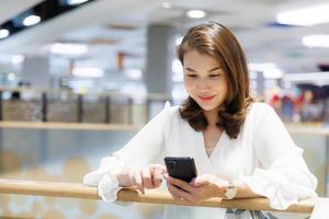 Beautiful Asian mature woman sitting indoors and using smartphone texting and sending while waiting for someone. Attractive female smiles and happy while looking at mobile phone screen photo