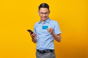 Portrait of smiling young asian man Asian in glasses holding mobile phone and credit card isolated on yellow background. businessman and entrepreneur concept photo