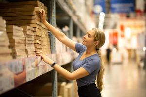 Cheerful woman customer looking up and pulling product on shelf while shopping in hardware store photo