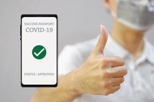 COVID-19 vaccine passport approved smartphone app with asian man wearing protective face mask selective focus thumb up background. Vaccination campaign and new normal prevention lifestyle concept. photo