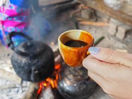 Close-up of hand woman holding a cup of coffee with old kettle on charcoal stove photo