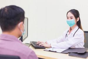 Professional Asian woman doctor records symptom of man patient in document at hospital both of them wear medical face mask to protect Coronavirus Covid 19 while in examination room at hospital. photo