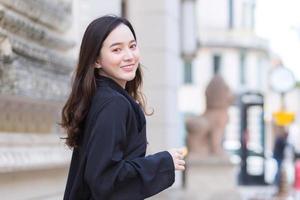 A picture of a beautiful long haired Asian beautiful woman in a black robe smiling happily walking and looking out in the city outdoors. photo