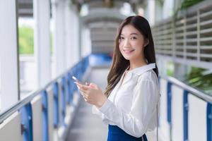 una mujer hermosa de negocios asiáticos se encuentra en un paso elevado de skytrain en la ciudad mientras usa su teléfono inteligente para enviar mensajes a alguien. foto