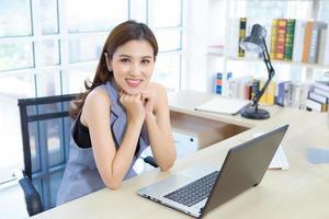 una hermosa mujer asiática profesional se sienta en una silla para trabajar sonriendo felizmente en la oficina, está mirando la cámara que tiene una computadora portátil y una lámpara puesta en la mesa. foto