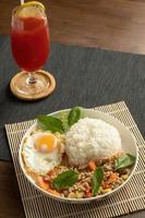 Stir-fried Thai basil with minced pork meat, carrots, and baby corns, with fried egg and fresh basils sort in a white dish on bamboo placemat, grey placemat, brown wood table. Copy space photo