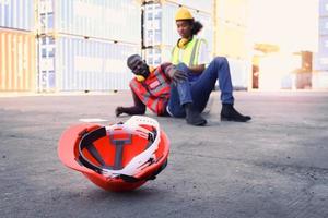 Safety helmet on ground with background of industrial African American engineer worker man getting hurt from accident at workplace, lying on floor at construction site area, colleague try to help him. photo