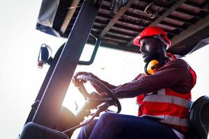 Industrial worker man wearing safety bright neon red vest and helmet driving forklift car at plant factory industry, African American engineer male working at logistic shipping cargo container yard. photo