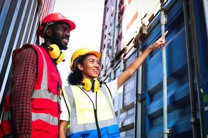 Two industrial African American engineer man and woman wear safety vest and helmet working together at logistic shipping cargo container yard, worker female pointing and asking opinion of colleague. photo