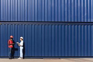 Two workers wear safety vest and helmet discus at logistic shipping cargo container yard workplace. African American engineer man talk with beautiful woman at blue containers background copy space.