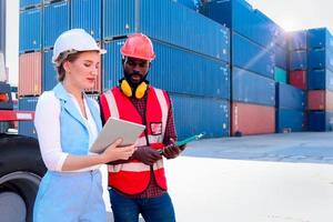 Two workers wearing safety vest and helmet discussing at logistic shipping cargo container yard. African American engineer man talking with beautiful young woman boss with blonde hair at workplace. photo