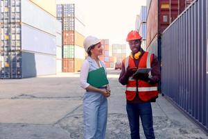 Two workers wearing safety vest and helmet discussing at logistic shipping cargo containers yard. African American engineer man talking with beautiful young woman boss with blonde hair at workplace.