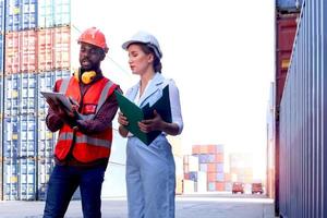 Two workers with safety helmet at logistic shipping cargo containers yard. African American engineer man using digital tablet and document folder for discussion with beautiful woman boss at workplace. photo