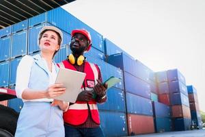 Two workers wearing safety vest and helmet discussing at logistic shipping cargo container yard. African American engineer man talking with beautiful young woman boss with blonde hair at workplace. photo