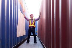 Portrait of senior elderly Asian worker engineer wearing safety vest and helmet, raising hand up, standing between red and blue containers background at logistic shipping cargo containers yard. photo