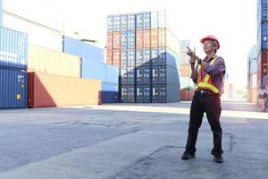 Senior elderly Asian worker engineer wearing safety vest and helmet, holding radio walkies talkie and pointing something at logistic shipping cargo containers yard. elderly people at workplace concept