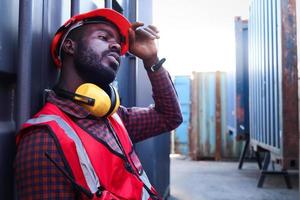 Portrait of African American young engineer worker man wears safety red color vest and helmet, feels tired from working hard, rests beside shipping container at logistic cargo yard. photo