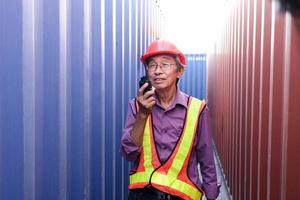 Portrait of senior elderly Asian worker engineer wearing safety vest and helmet, holding radio walkies talkie, standing between red and blue containers at logistic shipping cargo containers yard.