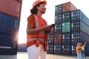 African American woman engineer worker with curly hair wearing safety vest and helmet, holding mobile phone at twilight sunset time at logistic shipping cargo container yard.