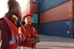 Happy smiling industrial African American engineer man and woman wear safety vest and helmet, two workers working at logistic shipping cargo container yard in twilight sunset time.
