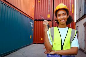Portrait of happy smiling beautiful African American woman engineer worker with curly hair wearing safety vest and helmet, holding walkies-talkie at logistic shipping cargo container yard.