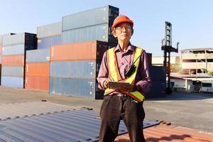 A senior elderly Asian worker engineer wearing safety vest and helmet standing and holding digital tablet at shipping cargo containers yard. elderly people at workplace concept photo
