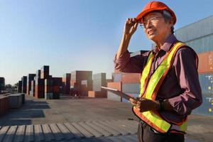 A senior elderly Asian worker engineer wearing safety vest and helmet standing and holding digital tablet at shipping cargo containers yard. elderly people at workplace concept photo