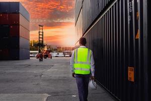 Behind industrial engineer worker wearing safety bright neon color vest and helmet, walking through logistic shipping cargo container yard workplace in twilight evening sunset time with orange sky. photo