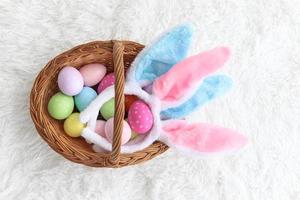 coloridos huevos de Pascua y diadema de orejas de conejo en una cesta de mimbre sobre una alfombra blanca esponjosa, decoración y celebración del comienzo de la primavera. foto