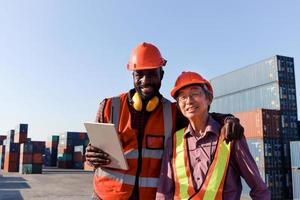 Two workers wear safety helmet work together at logistic shipping cargo containers yard. African American and senior elderly Asian engineer hug each other, respect in differences of race skin color. photo