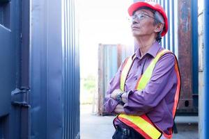 Portrait of serious senior elderly Asian worker engineer wearing safety vest and helmet, standing with arms crossed with blue containers as background at logistic shipping cargo containers yard.