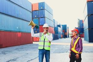Two workers with safety vest and helmet working at logistic shipping cargo containers yard, the engineer boss holding megaphone speaker for communicating with serious senior elderly Asian staff. photo