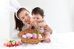 Happy smiling adorable six months baby girl playing with wicker basket of colorful Easter eggs in embrace of mother arms, mom with rabbit ears headband holding her sweet little daughter kid.