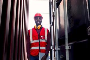 trabajador ingeniero afroamericano con chaleco y casco de seguridad de color rojo neón brillante, sosteniendo una tableta digital y parado entre contenedores en el patio de contenedores de carga de envío logístico. foto