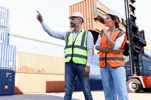Portrait of two workers wearing safety vest and helmet discuss at logistic shipping cargo container yard, senior engineer man point and ask opinion of beautiful young woman colleague at workplace. photo