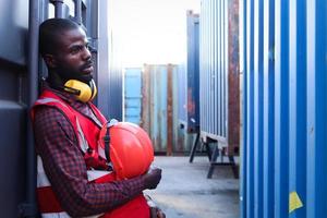 Portrait of African American young engineer worker man with safety red color vest and helmet, feeling tired from working hard, resting beside shipping container at logistic cargo yard. photo