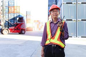 A senior elderly Asian worker engineer wearing safety vest and helmet standing and holding radio walkies talkie at logistic shipping cargo containers yard. elderly people at workplace concept. photo