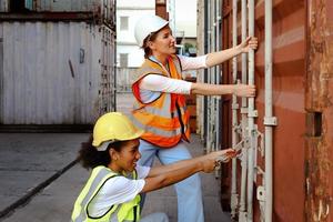 dos hermosas jóvenes trabajadoras ingenieras usan chaleco y casco de seguridad, intentan abrir la puerta del contenedor de envío en el patio de carga logística. las niñas afroamericanas y caucásicas trabajan juntas en el mismo lugar de trabajo. foto