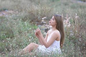 hermosa mujer joven sentada en el campo de hierba verde y soplando diente de león. al aire libre. disfruta de la naturaleza. niña sonriente saludable en el césped de primavera. concepto libre de alergias. libertad foto