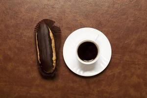 black coffee, chocolate Eclair, coffee in white Cup, white saucer, on brown table, Eclair on stand