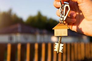 Key ring with keychain house in hand. Background of fence and cottage. Moving to a new home, mortgage, buying real estate, renting and booking housing, dream of living  in the village. Copy space photo