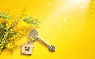 Key ring in the shape of wooden house with key on a yellow background and spring mimosa. Building, design, project, moving to new home, mortgage, rent and purchase real estate. Copy space photo